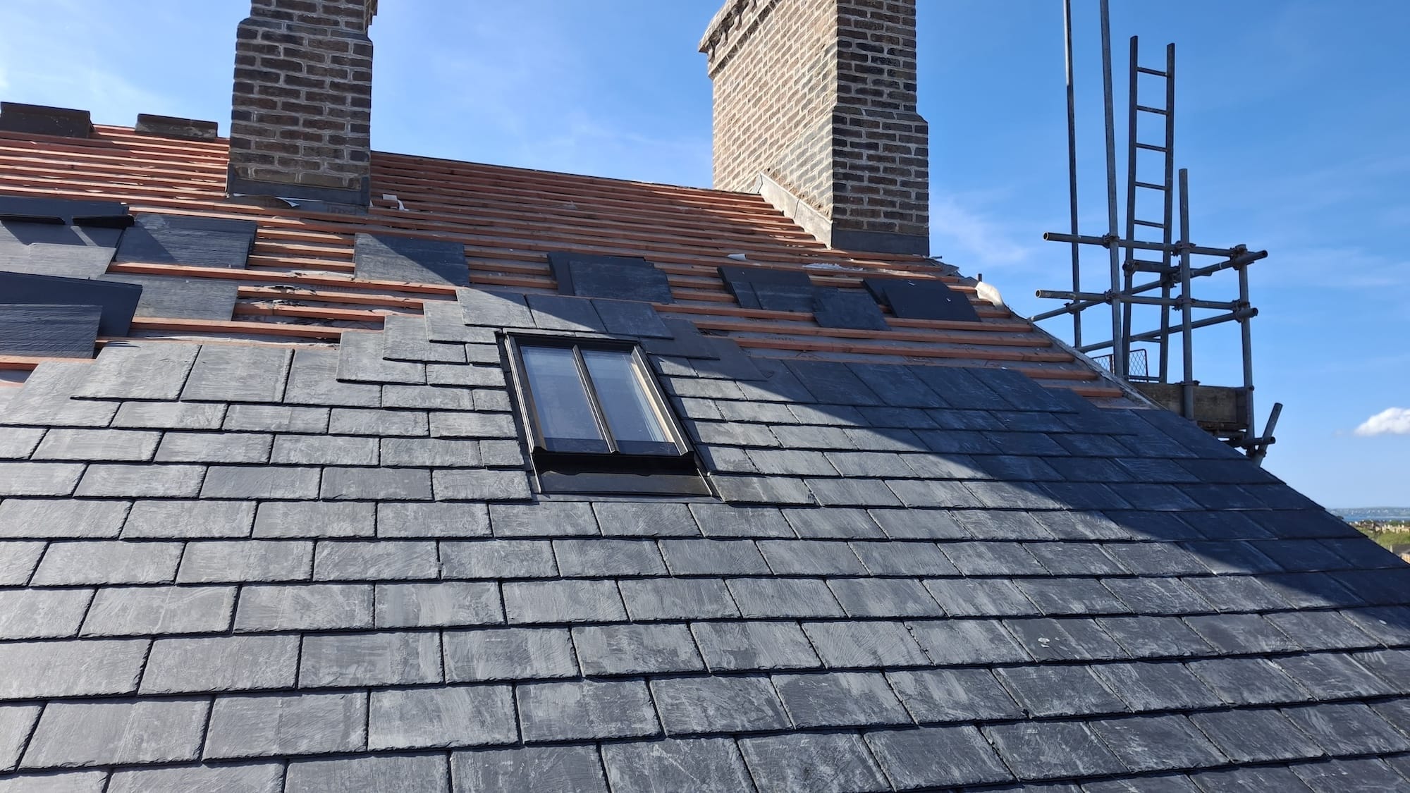 A partially completed slate roof with a window, two chimneys, visible wooden battens, and scaffolding on the right side under a clear blue sky during the architectural renovation of Y Tŵr community hotel.