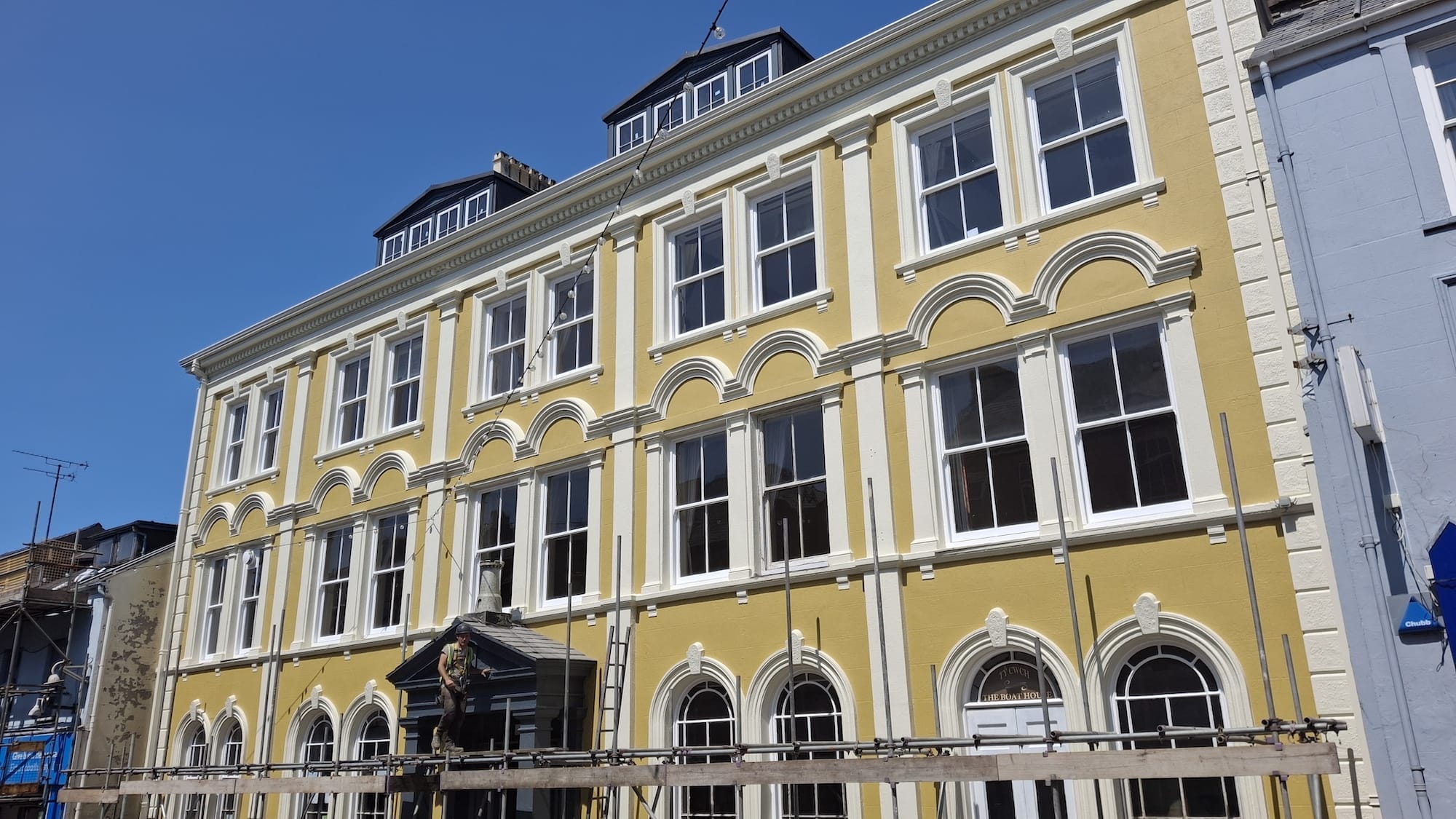 A yellow three-story building with white trim and arched windows, viewed from below on a sunny day, stands amid scaffolding and a ladder during the architectural renovation of Y Tŵr community hotel.